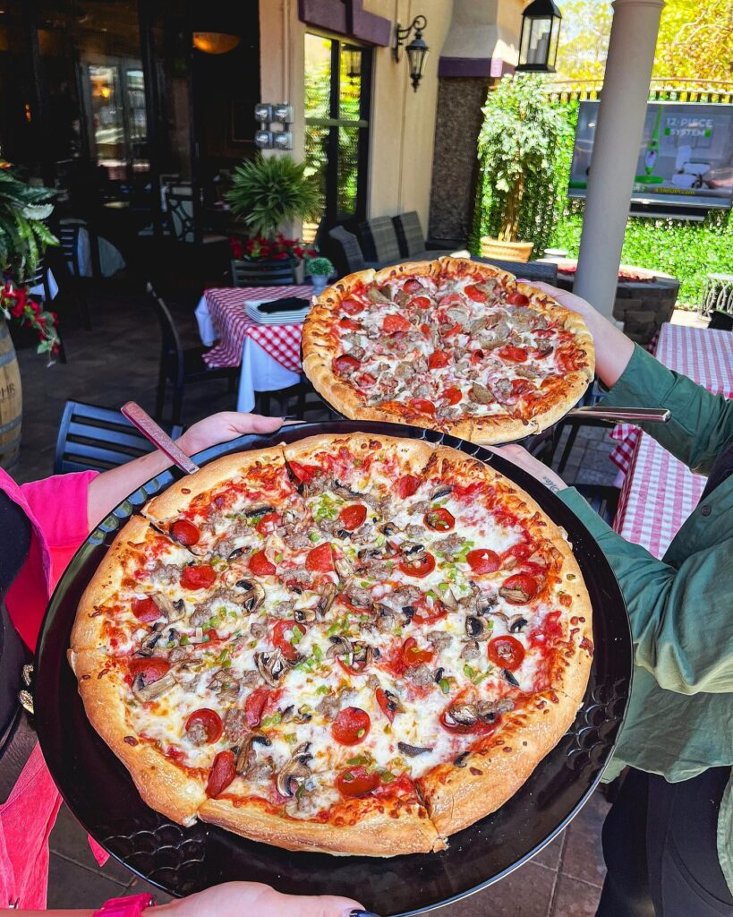 Two people holding two large pizzas with various toppings on a patio with red-checkered tablecloths and plants in the background.